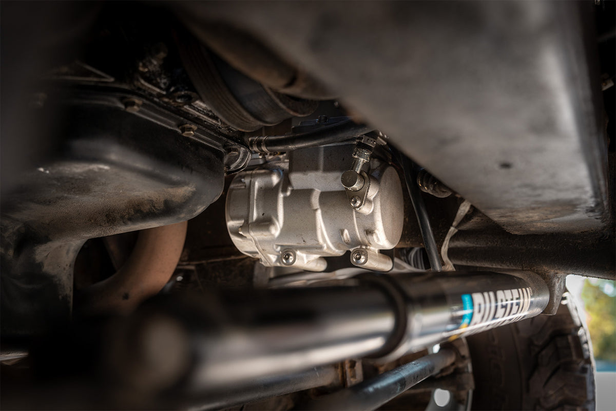 Close-up of a silver metal AC compressor and suspension components under a vehicle, with a visible Shock brand shock absorber and dark mechanical surroundings.