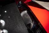 Close-up of a black metal motorcycle exhaust component with an embossed logo, set against a bright red painted body panel and a textured black surface.