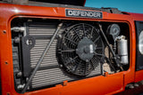 Close-up of an orange Defender vehicle front grille and radiator area, showing a black cooling fan, silver radiator, and a Defender badge above the grille.